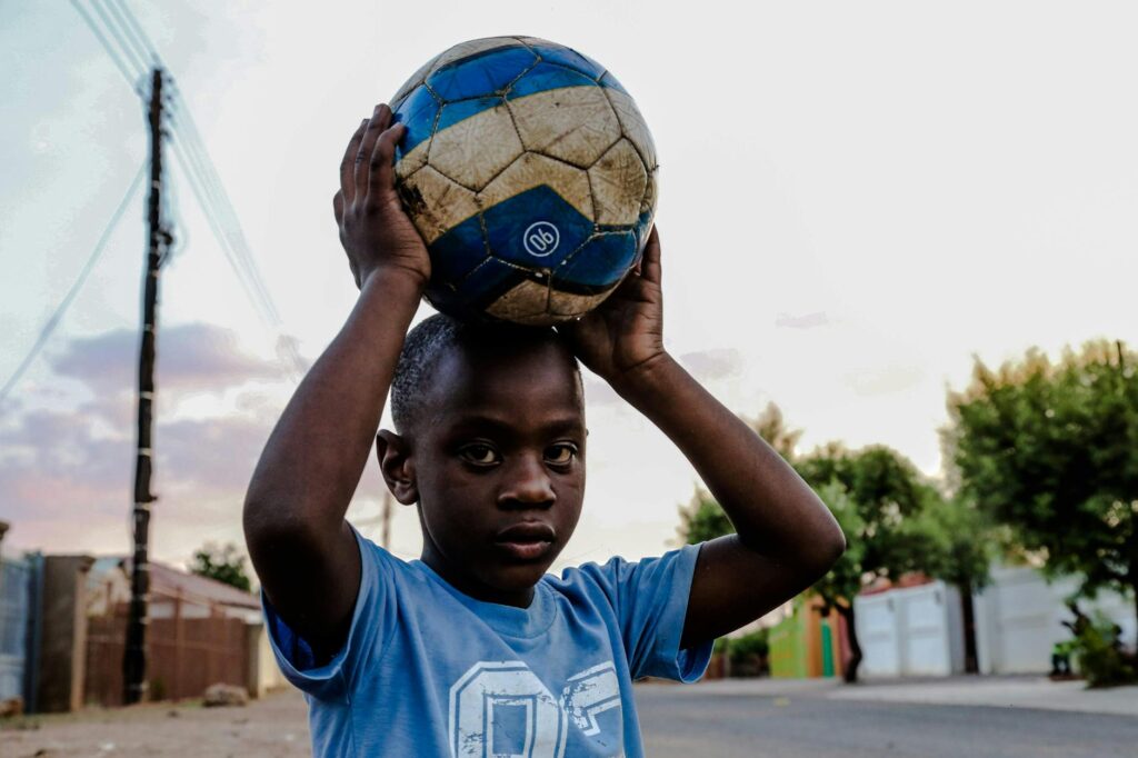A young boy in a blue shirt holds a soccer ball on a sunny day, standing on an urban street.