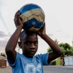 A young boy in a blue shirt holds a soccer ball on a sunny day, standing on an urban street.