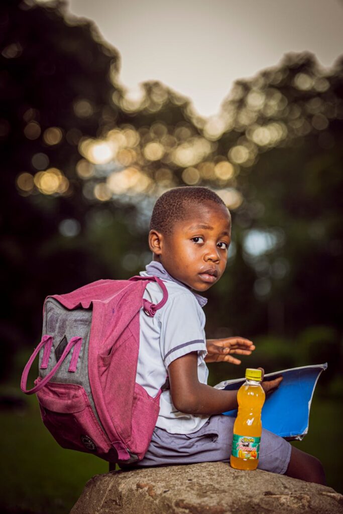 Young boy sitting outside with backpack, notebook, and juice bottle.